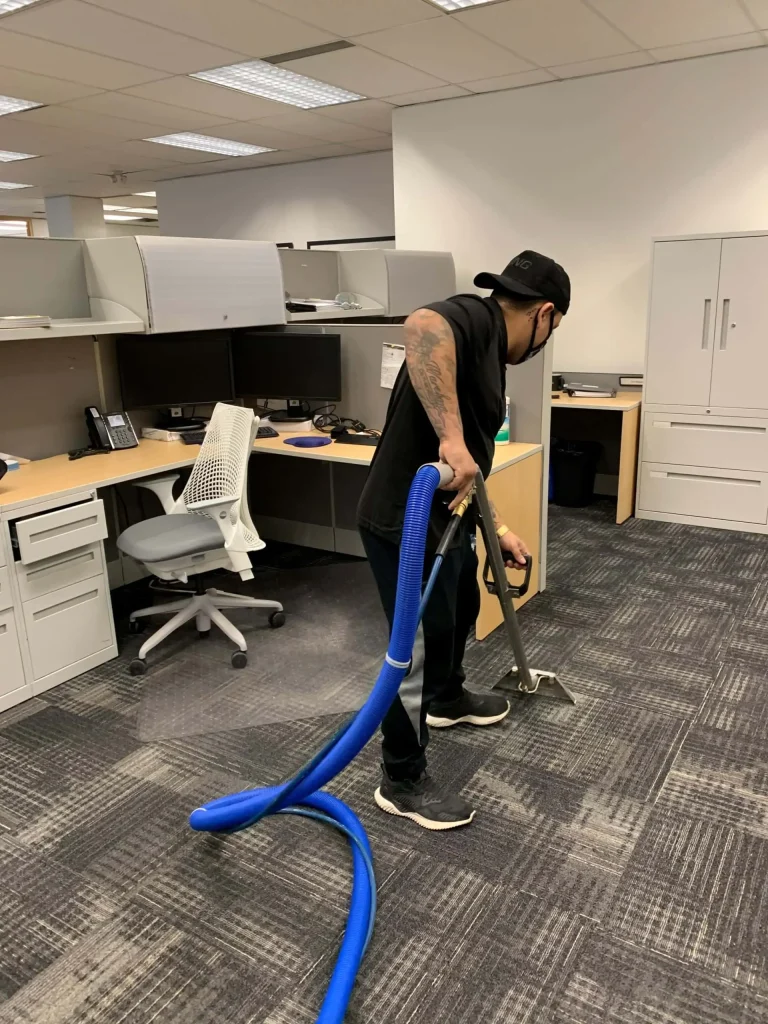 A male office cleaner in a black cap and sleeveless shirt vacuuming the carpet between office cubicle workstations with a blue vacuum hose