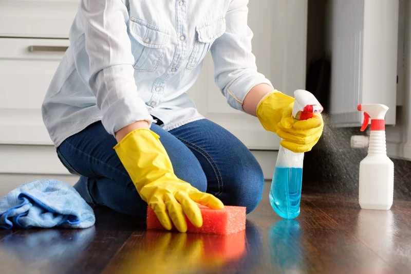 A cleaner in yellow rubber gloves kneeling on a floor spraying blue cleaning solution and scrubbing with a red sponge next to a microfiber cloth and spray bottle