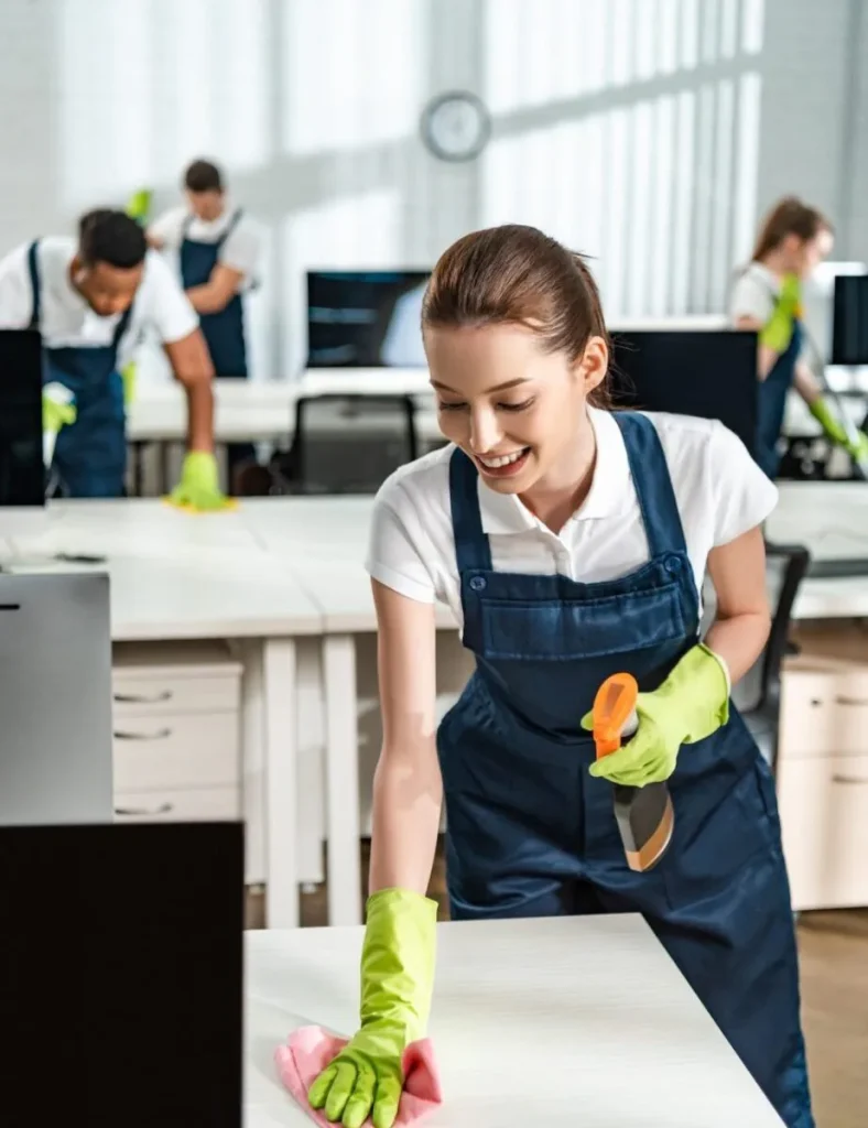 A smiling female cleaning professional in navy overalls and green rubber gloves wiping a white countertop with a spray bottle in hand